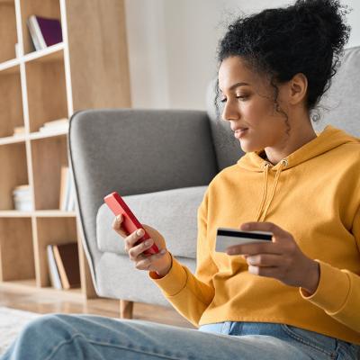 A young woman sitting on the floor of a living room, holding a credit card and browsing her smartphone.