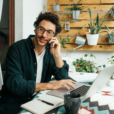 A young man on phone call and using laptop to work remotely from home.