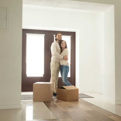 A hugging couple stand in the house in front of the door with two cardboard boxes in front of them.