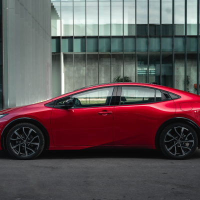A red 2024 Toyota Prius Prime parked in front of a building on a city street.