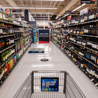 A shopping cart in a grocery store aisle filled with wine bottles.