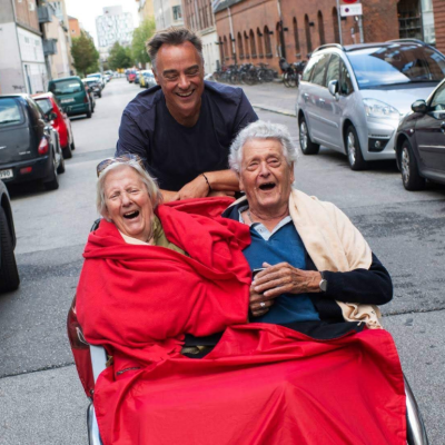 A volunteer cycles with a trishaw carrying two seated passengers who are laughing and smiling. 