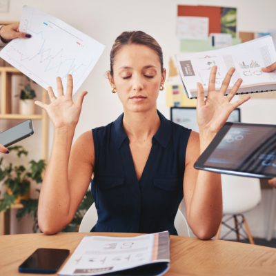 A women sitting in an office closes her eyes and puts her hands up as people hand her things including a phone, a computer, a paper with a graph on it, and a project notebook.