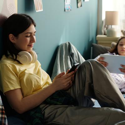 Two teenage girls relaxing on a bed and using smart devices to browse the internet.