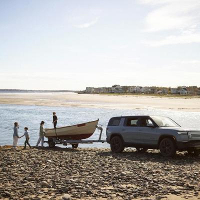 Rivian R1S towing a boat, parked near a body of water on a rocky dirt beach. A family of four plays near the boat.
