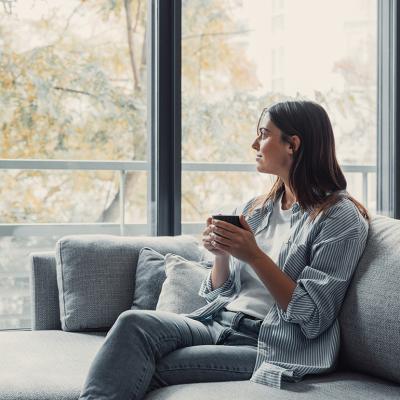 Young woman sitting on a sofa and having a cup of coffee while looking out the window at home.
