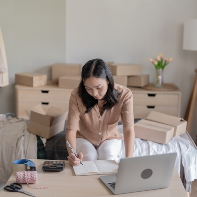 A woman writing in a notebook on a desk with a calculator, a laptop, scissors, tape and string, in a room with clothes hanging on a rack and cardboard boxes stacked on various surfaces.