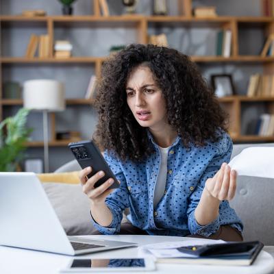 A young woman at home stressfully looking at her smartphone while calculating expenses.