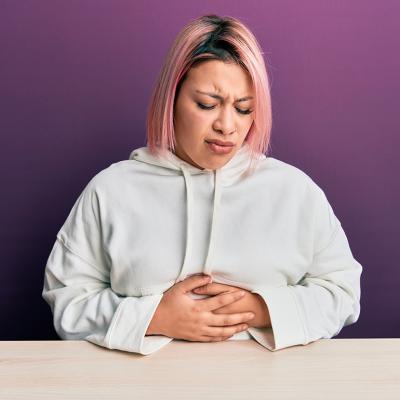 A woman with pink hair sitting with her hands on her stomach, feeling unwell.