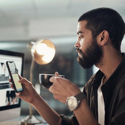 A man in a creative office checking social media on his phone while drinking a cup of coffee.