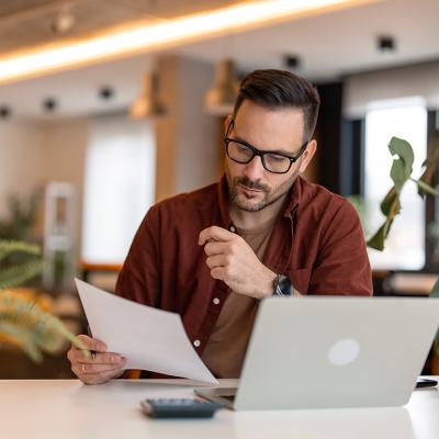 A person looking at a paper and using a laptop in a home office. 