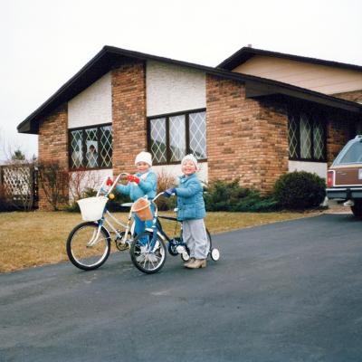 A vintage photo of two 80s siblings, boy and girl, riding bikes in their driveway.