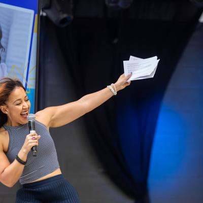 Julia Collins who is the founder and CEO of Planet FWD, speaks onstage with one hand holding a microphone and the other arm raised, her portrait is visible on a screen in the background.