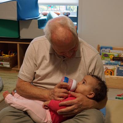 Teacher Herb Hickey holds a baby while she drinks from a bottle.