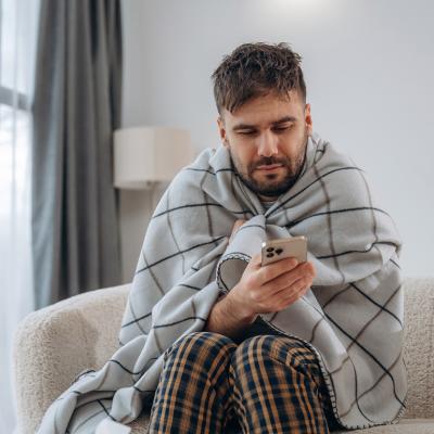 A sick man at home wrapped in blanket and looking up information on his phone.