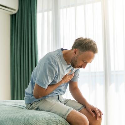 A man sitting on his bed clutching chest due to pain.