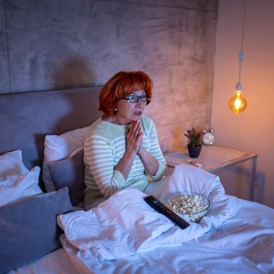 A senior woman watching a scary movie on the TV from bed.