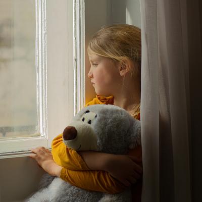 A kid with a stuff toy looking out in a window. 