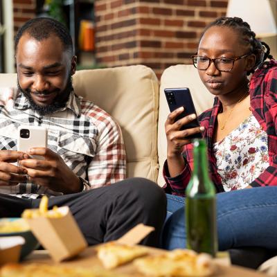 A black couple relaxing on a sofa at home and individually using their smartphones.