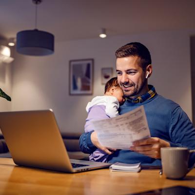 A young father carrying his baby asleep while working remotely from home.