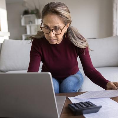 A focused senior woman holding financial documents and browsing information on a laptop.