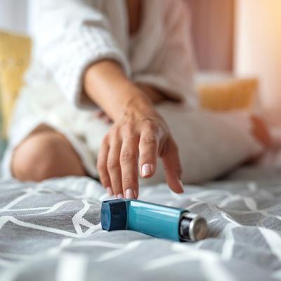 Woman sitting on a bed reaching for her inhaler.