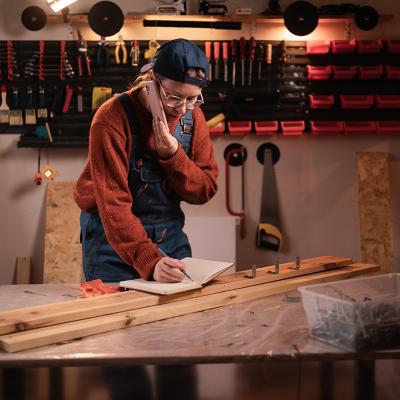A female carpenter writing down notes while talking on the phone in a workshop.