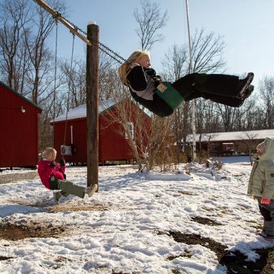 Two children on a swingset while another child watches on a snowy day outside Nature's Gift Microschool.