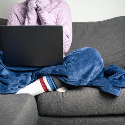 Person wearing a knitted sweater and socks with a blanket on while working and sitting on a sofa.