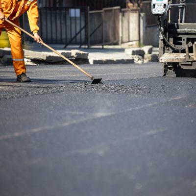 A construction worker in an orange uniform spreads freshly poured asphalt on a road surface.