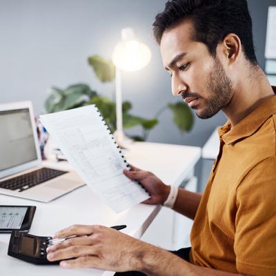 A finance analyst reviewing financial documents using a calculator and a work laptop in an office.