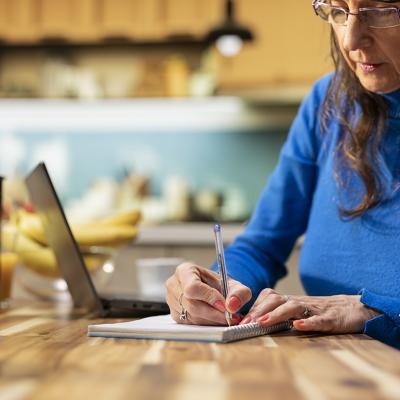 An elderly person writing a grocery list in a kitchen table.