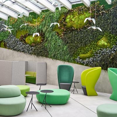 A modern office interior featuring a large biophilic living wall covered in various mosses, ferns, and plants spanning the back wall beneath a glass skylight ceiling with white beams. The space includes contemporary furniture in shades of green and lime.
