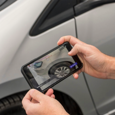 Two hands holding a smart phone aimed at a silver car, the phone's screen shows an AI-powered scan of possible vehicle damage.