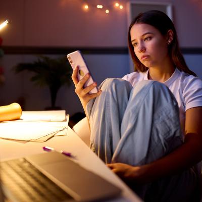 A young woman sitting at a desk in her bedroom and browsing her phone at night.