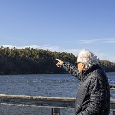Protect Wiscasset co-founder Richard Davis facing away from the camera wearing a black jacket and his white air blown by the wind, points to the undeveloped land across a waterway where the prospect of a $5 billion data center emerged.