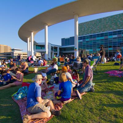 Groups of people sit on grass as they wait for an outdoor concert at the Long Center Lawn in Austin, Texas.