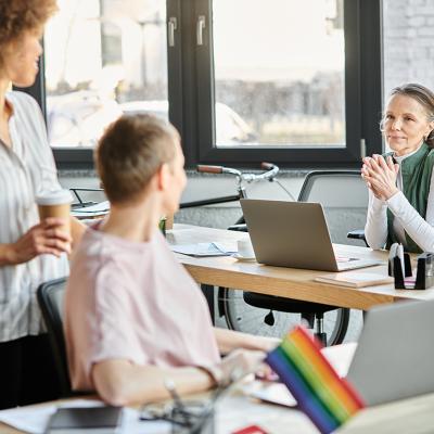Business partners collaborating on a project and a rainbow flag seen from a desk indicating it as a LGBTQ+ inclusive workplace. 