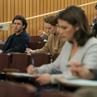 A publicity still from the first season of the TV show Off-Campus shows two people in the same college lecture with the focus on one man watching another woman while seated in the row behind her.