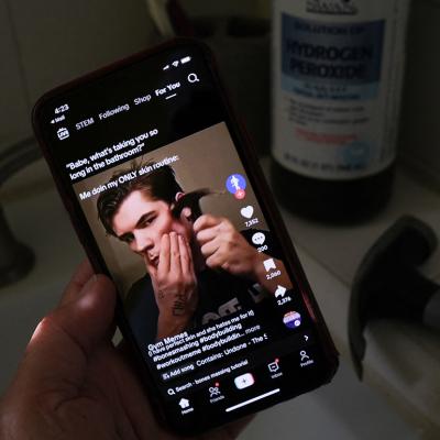 A person watching from his smartphone a video of a male TikTok influencer hammering his cheekbone, while a hydrogen peroxide bottle and a hammer are also seen in his bathroom.