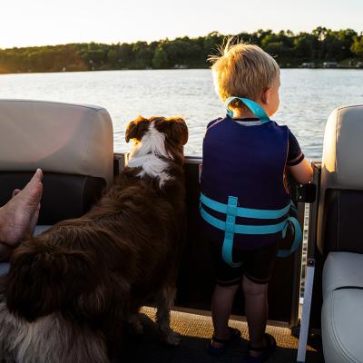 A toddler wearing a life vest and their family dog looking at the waters from a boat. 