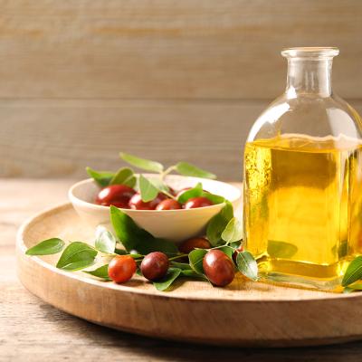 A glass bottle of jojoba oil with leaves and seeds of a shrub in a wooden plate.