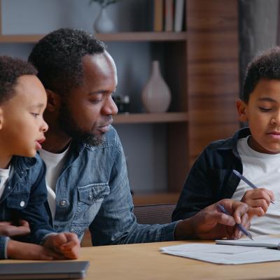 A parent studying with his young children, while helping his other son with a school writing assignment.
