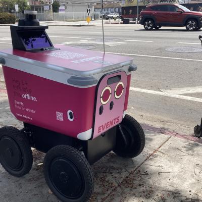 A pink sidewalk delivery robot built by California-based Serve Robotics features an ad from dating app Tinder, while sitting on a sidewalk next to a shopping cart with cars on the road in the background.