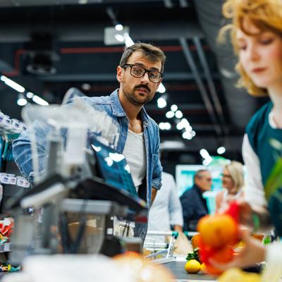 A male customer wearing glasses and denim shirt watches the cashier scanning groceries at a checkout.