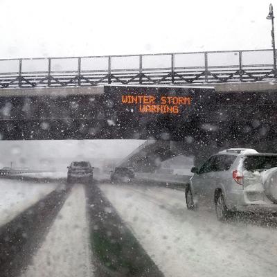 Windshield POV of a car passing under a bridge with an electronic 'Winter Storm Warning' sign.