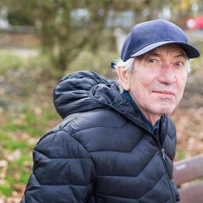 An elderly man wearing a thick jacket and hat while sitting alone on a bench in a park.