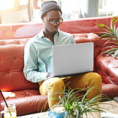 A fashionable young black freelancer working remotely on a laptop while sitting on a red leather sofa.
