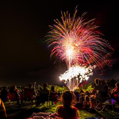 Crowds of people sit in lawn chairs and blankets to enjoy the colorful fourth of July independence day celebratory fireworks exploding in the black night sky.