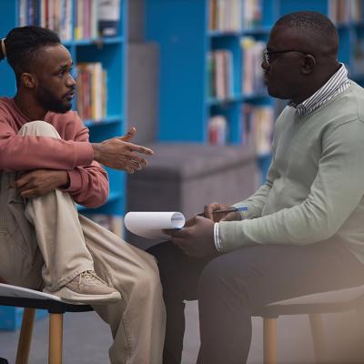 A young black male student talking to a counselor in their college library.